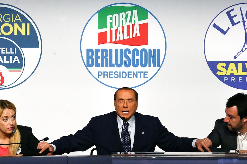Silvio Berlusconi speaks flanked by Giorgia Meloni (left) and Matteo Salvini (right) on stage.