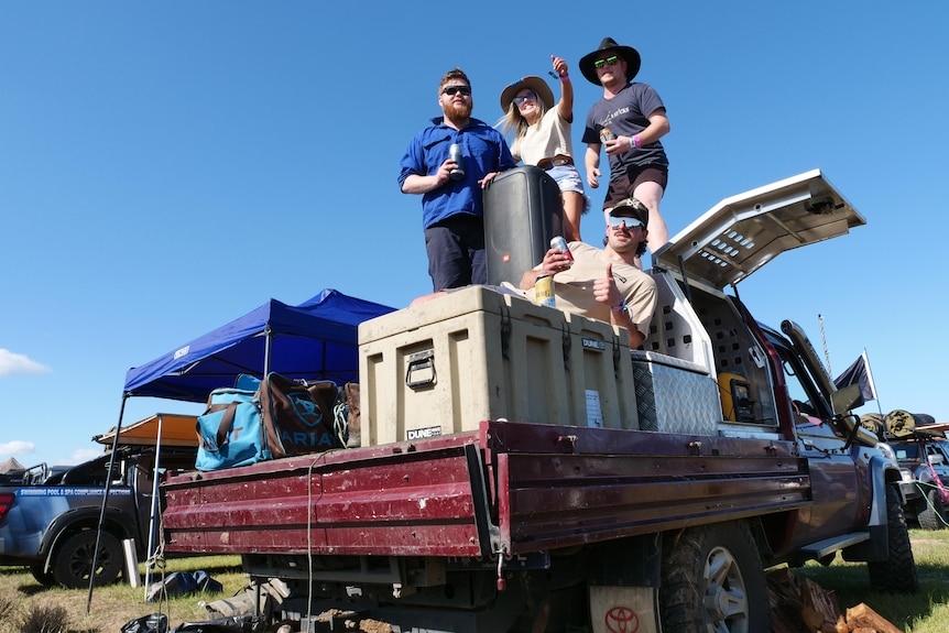 Three men and a woman pose on the back of a ute, with large eskies and speakers in the tray.