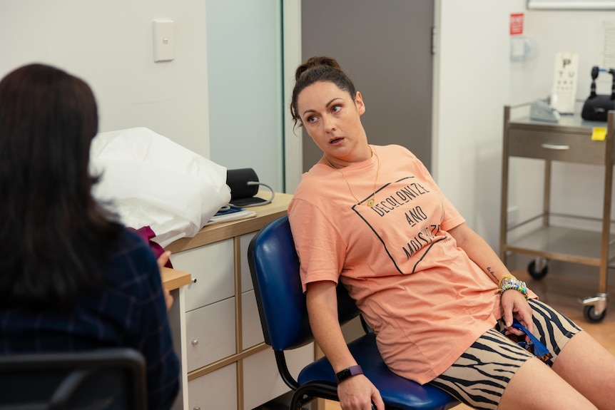 Une femme dans la quarantaine - la comédienne Celeste Barber - est assise affalée sur une chaise dans un cabinet médical dans la série télévisée Wellmania