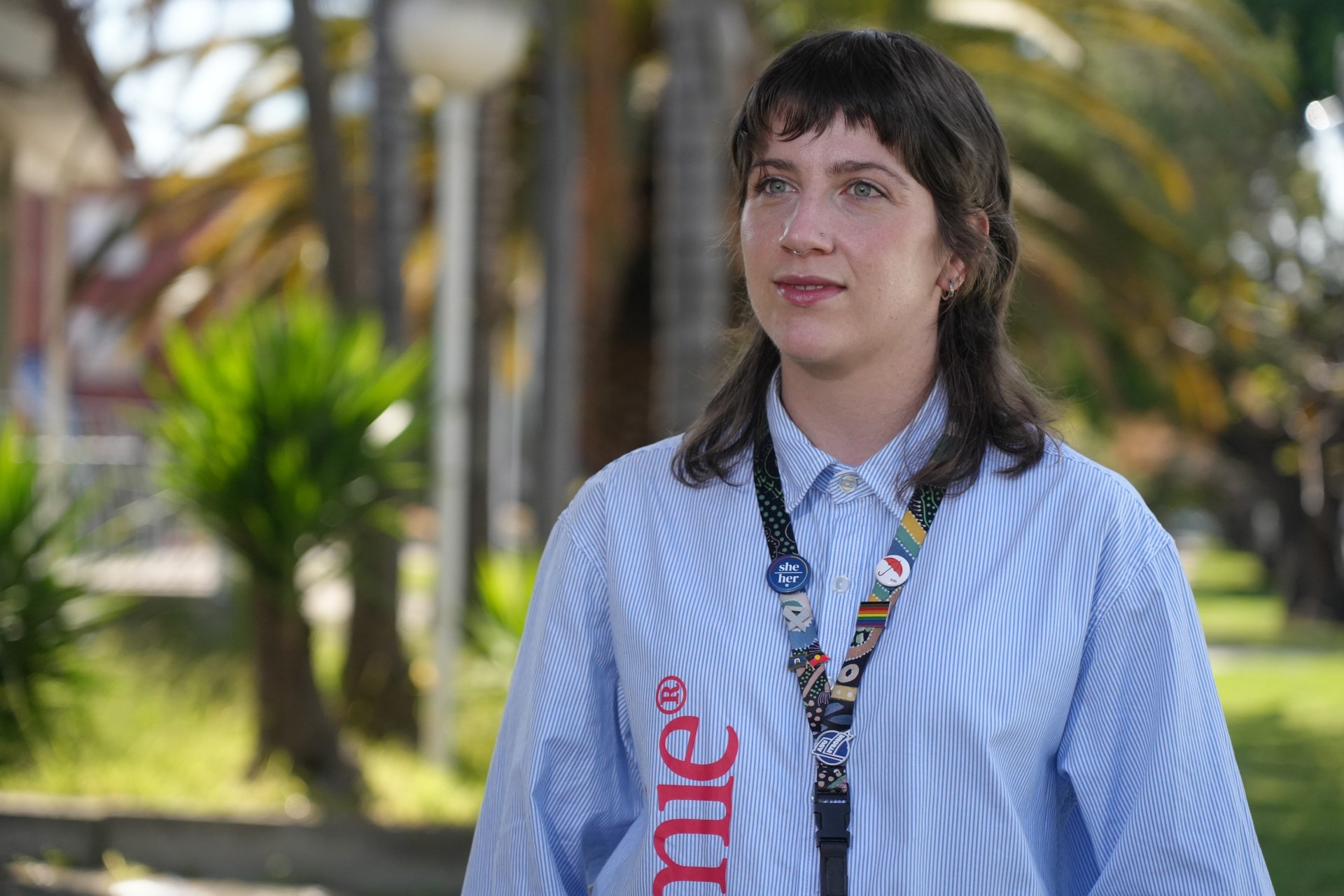 Georgia Harraway-Jones wears a pale blue, long-sleeved collared shirt. She is standing outside with trees behind her.