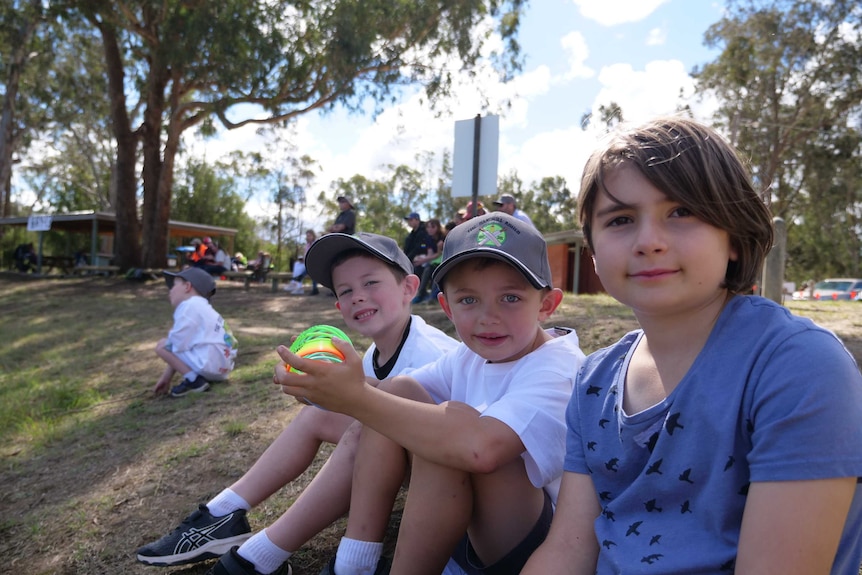 Nina Felmingham, Lochie Strecker and Charlie Henderson watching cricket at a country oval surrounded by trees.