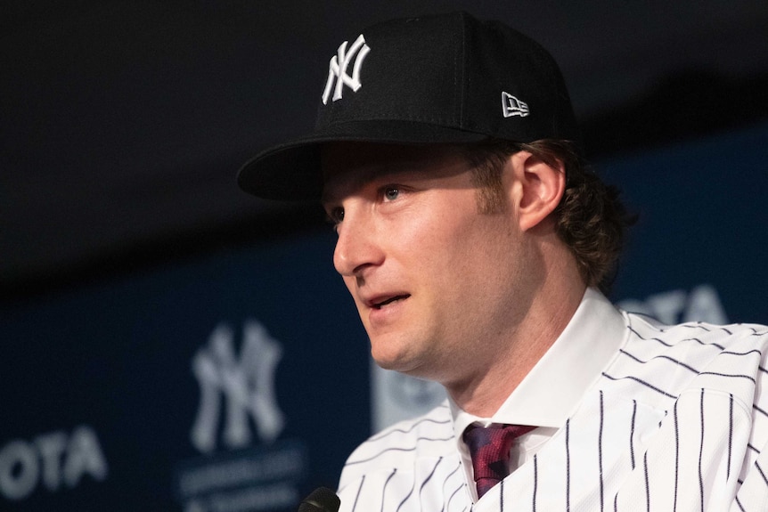 Gerrit Cole wears a pin striped shirt over a conventional shirt and tie, with a Yankees hat