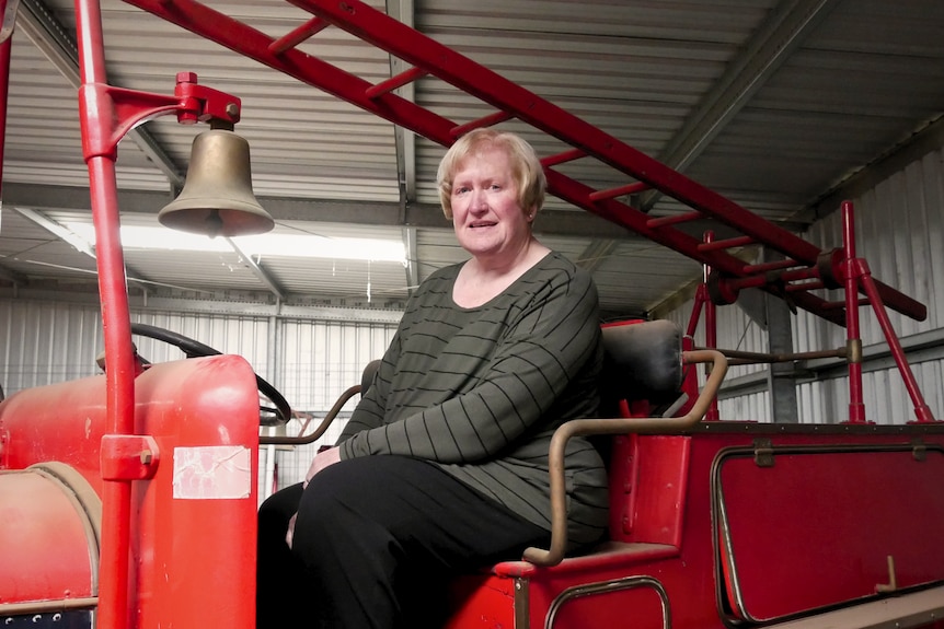 A woman sits on an old red fire engine with a brass bell and ladder overhead.