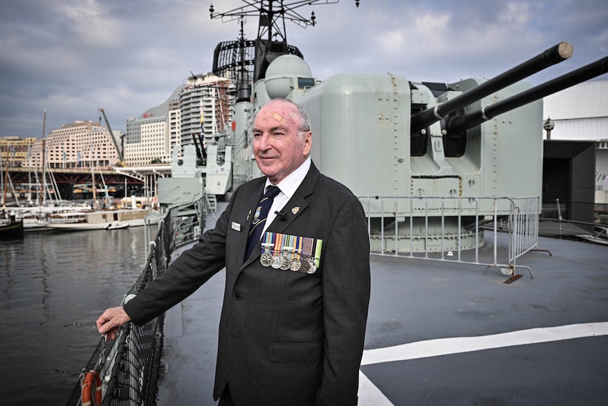 An older man in military colours stands on a destroyer near the turrets