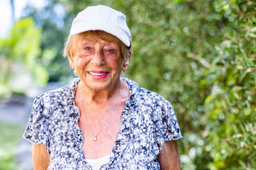 An elderly woman in a hat and floral shirt stands smiling beside a hedge.