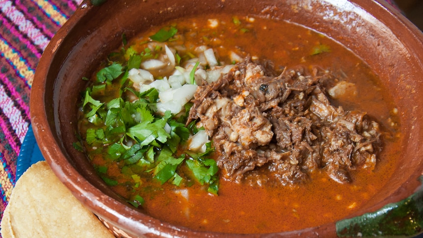 Juicy beef consomme topped with onion, coriander and pulled beef, set in clay dish with tortillas and cut lime.