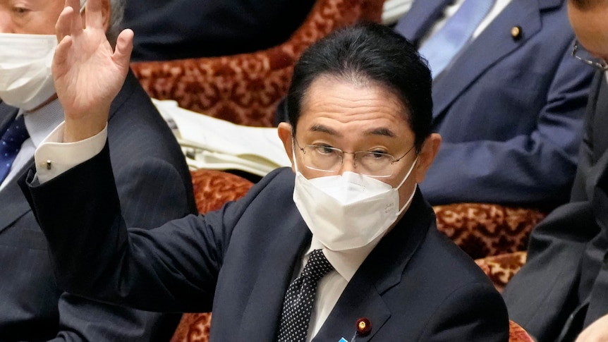 Japanese PM Fumio Kishida wearing a suit and face mask raises his hand. 