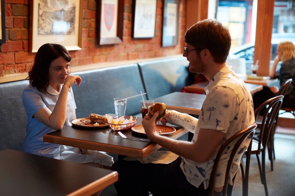 Emily Barclay sits left in a booth and looks at Tom Ward sat opposite her and who holds a burger mid-conversation. 