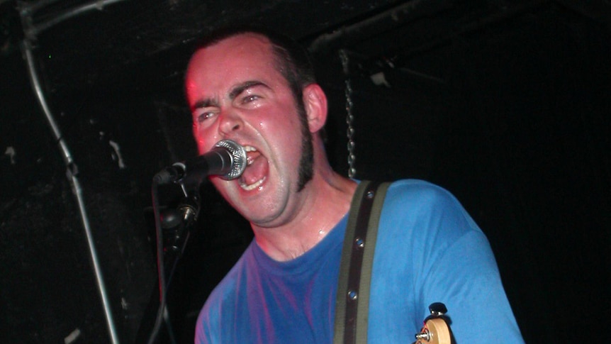 A sweaty Andy Falkous wearing a blue shirt screams into a microphone on stage