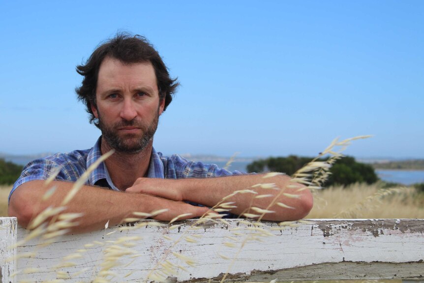 A man leans on a white wooden gate looks sadly at the camera with a water mass in the background.