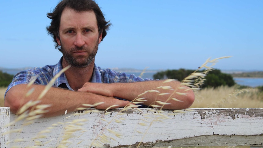 A man leans on a white wooden gate looks sadly at the camera with a water mass in the background.