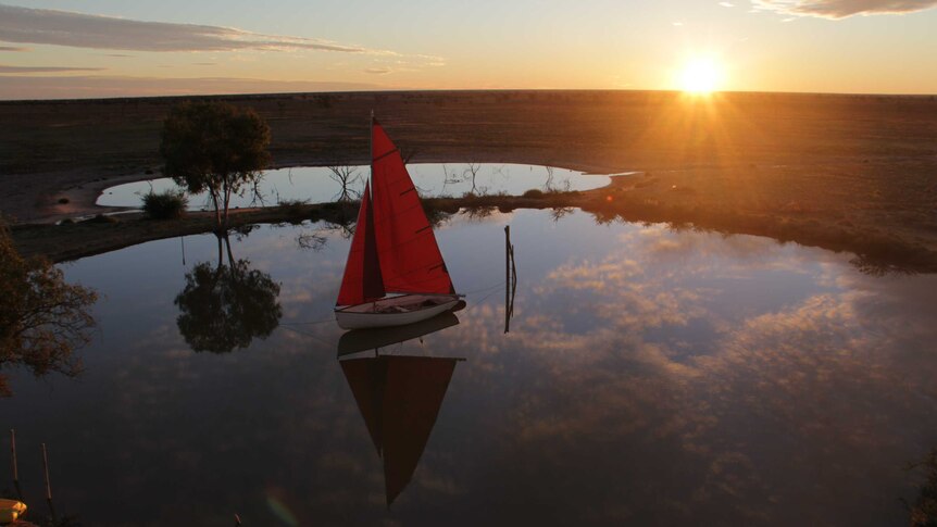 A boat with a red sail sits on a dam with the sun setting in the background.