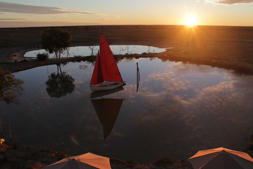 A boat with a red sail sits on a dam with the sun setting in the background.