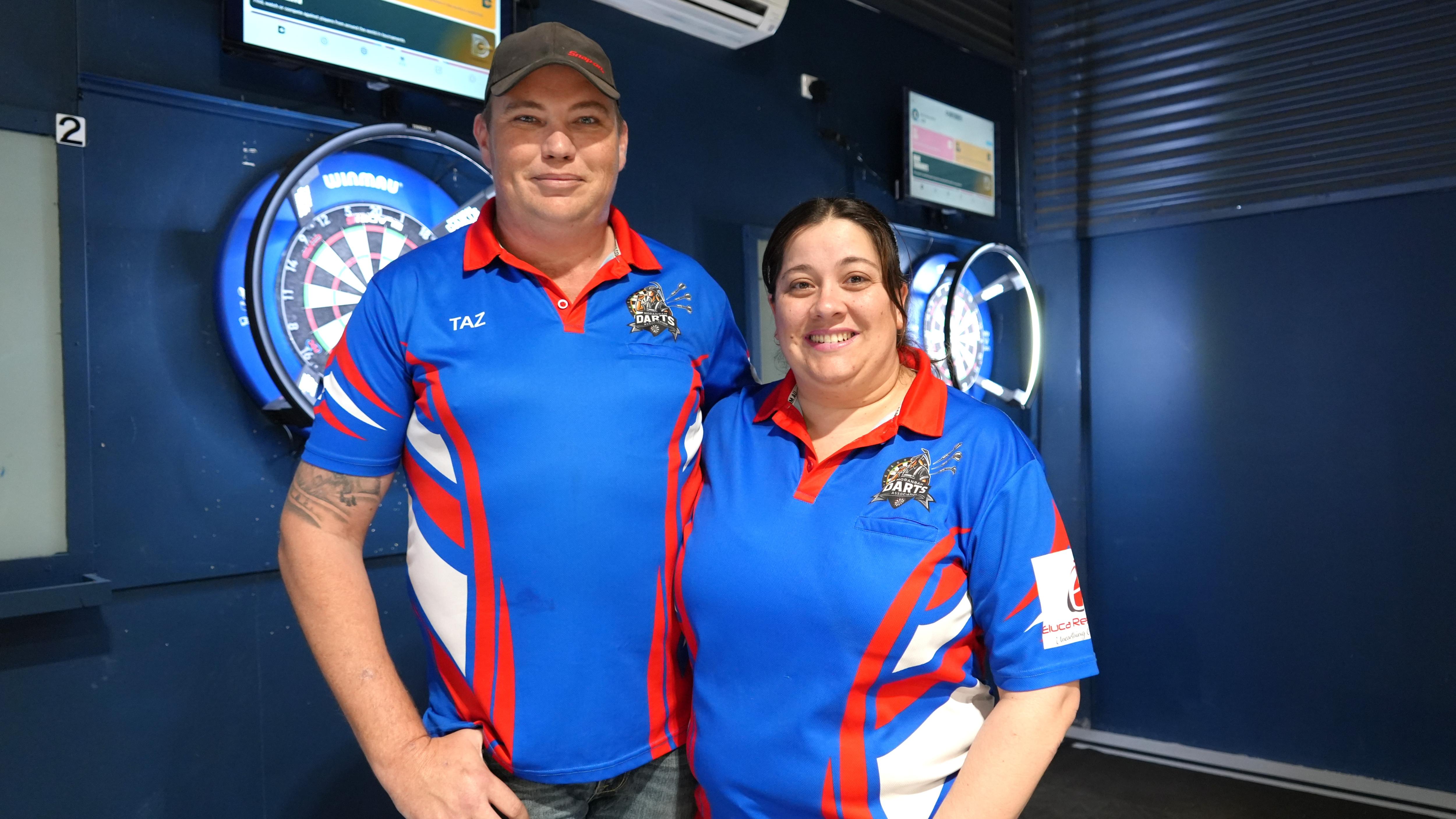 Man and woman in blue and red graphic sports shirts in front of darts board in hall
