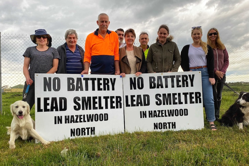 A group of community members stand outside the site of a proposed battery recycling centre with two signs.