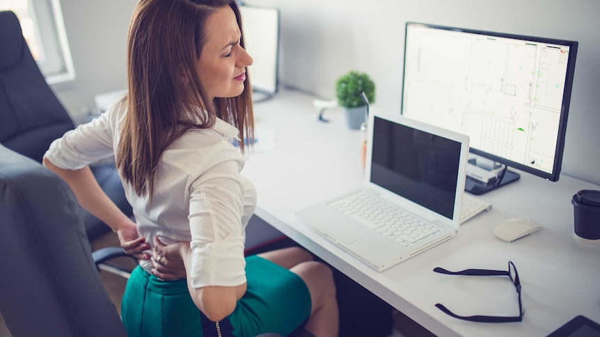 A woman sits at her office desk, holding her lower back in pain.