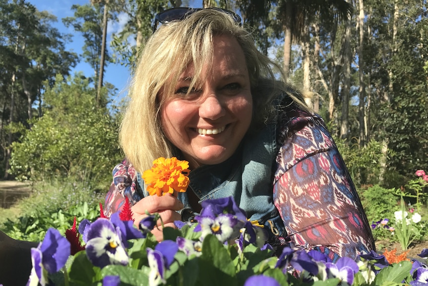 Sharon Koski crouches down behind a flower bed, smiling at the camera with a yellow marigold in her hand.
