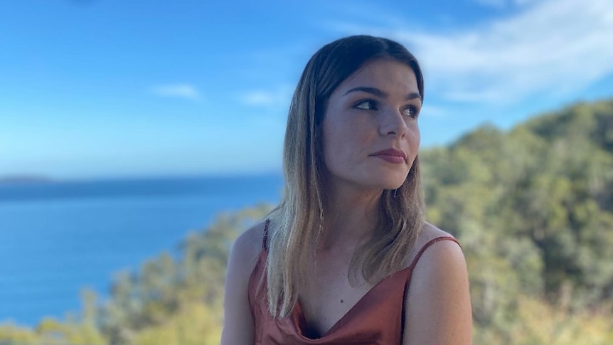 Grace wears a rust coloured cowl neck dress and looks to the right while standing in front of the sea and mountains.