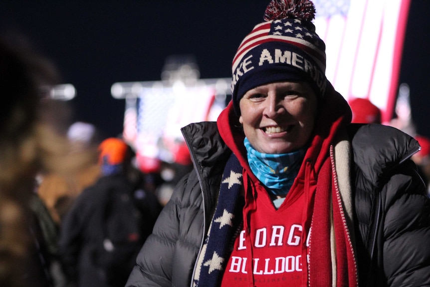 A woman wearing a make america great again beanie and puffer jacket smiles.