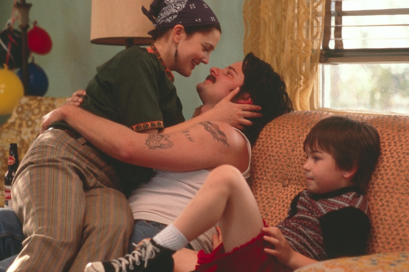 A young woman wearing bandana on head sits in loving embrace with man with tattooed arms on 70s style couch near a child.