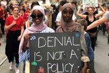 Two women hold a sign saying 'denial is not a policy' as they walk through a street crowded with protesters.