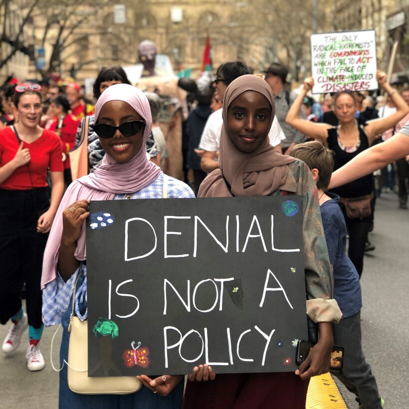 Two women hold a sign saying 'denial is not a policy' as they walk through a street crowded with protesters.