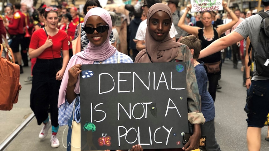 Two women hold a sign saying 'denial is not a policy' as they walk through a street crowded with protesters.