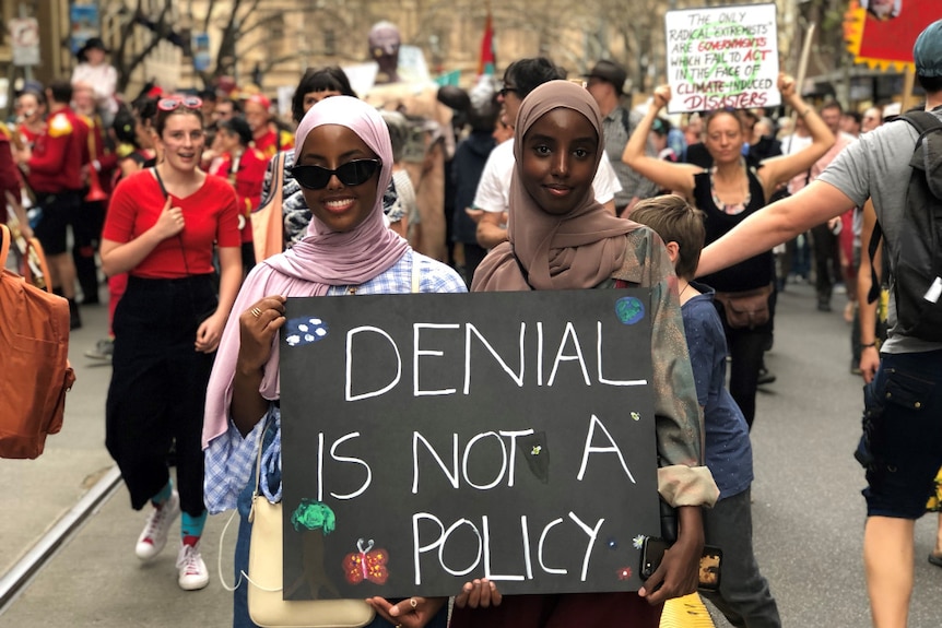 Two women hold a sign saying 'denial is not a policy' as they walk through a street crowded with protesters.