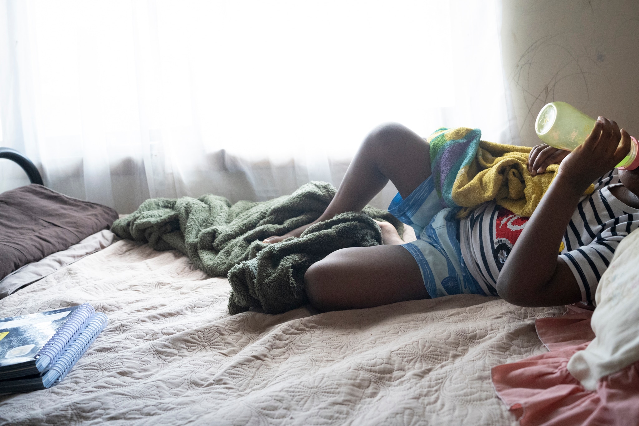 A child lies on a bed, their head can't be seen and they are drinking from a drink bottle.