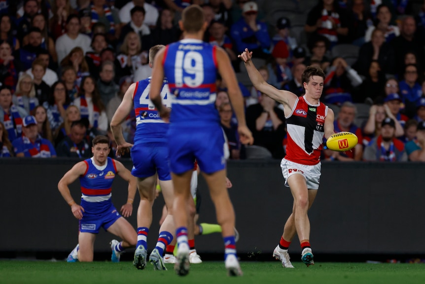 Western Bulldogs AFL defenders stand watching while a St Kilda forward holds the ball to snap a kick for goal around the corner.