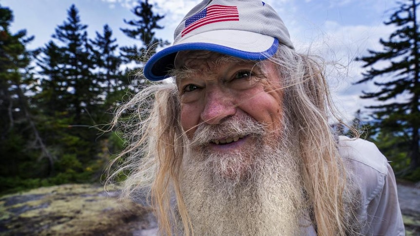 A man with long white beard and grey hair leans into the camera smiling, among pine trees outside 