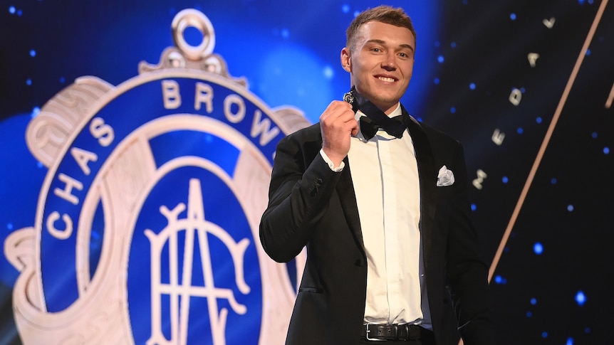 A Carlton AFL player stands on stage with a medal around his neck, and a sign behind him with the image of the Brownlow Medal.