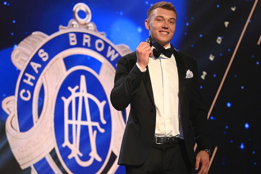 A Carlton AFL player stands on stage with a medal around his neck, and a sign behind him with the image of the Brownlow Medal.