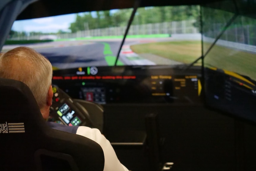 A man in a white shirt with white and grey hair driving in a Ferrari simulator