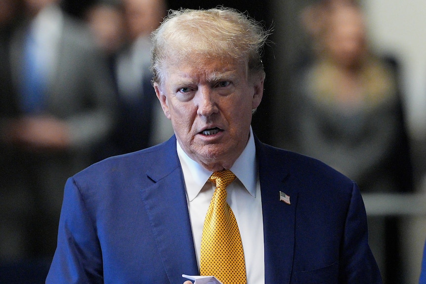 Donald Trump steht im blauen Anzug, mit gelber Krawatte und weißem Hemd da und spricht außer Sichtweite mit Reportern und sieht ernst aus