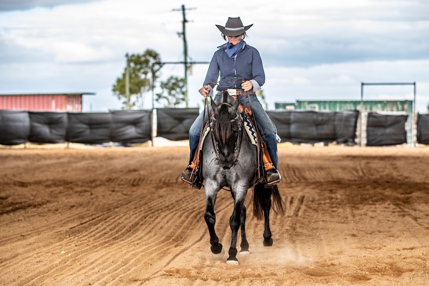 a woman in a navy button down shirt and have rides a grey horse on a dirt track