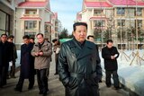 Kim Jong Un in a black leather trench coat, with his hands in his pockets, walking through a snowy streets with men behind him