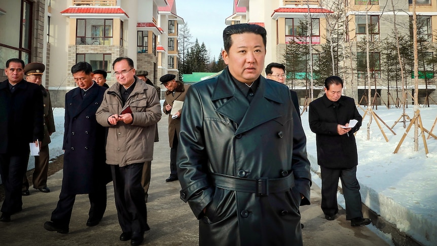 Kim Jong Un in a black leather trench coat, with his hands in his pockets, walking through a snowy streets with men behind him