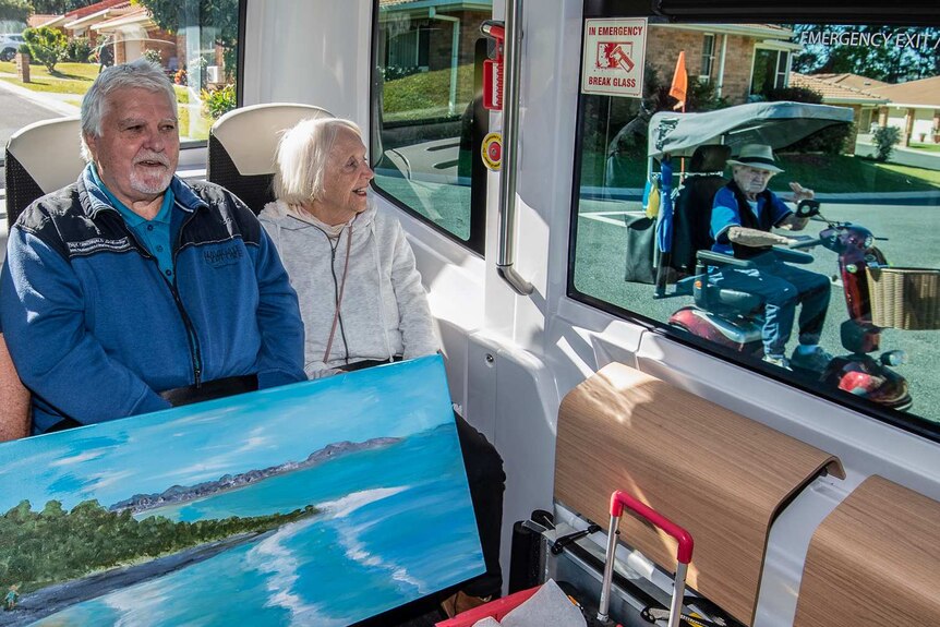 Three passengers sit on the bus while a resident in a scooter waves through the window.