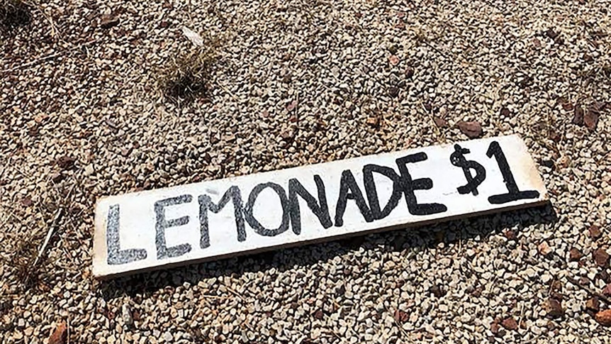 A photo of a sign advertising $1 cups of lemonade left on gravel bitumen.