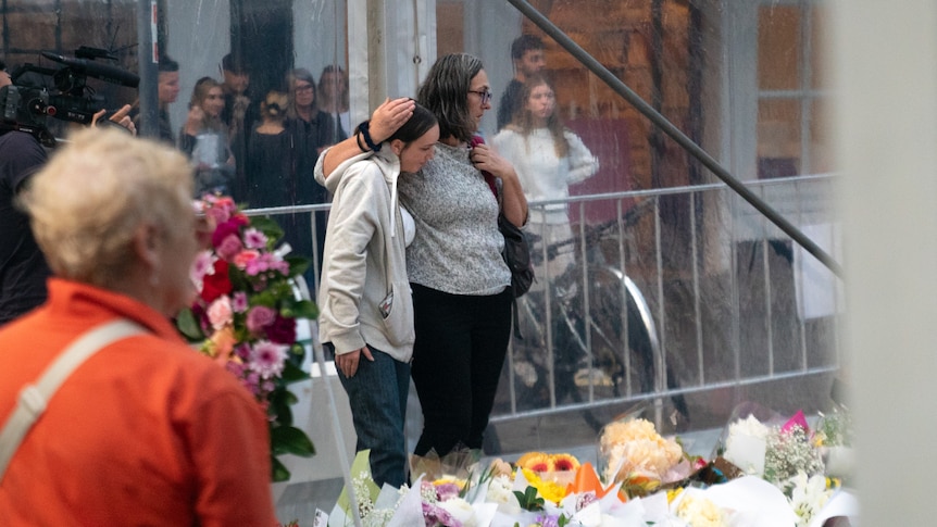 One woman stands with her hands around another womans shoulder looking at a sea of floral tributes on a street.
