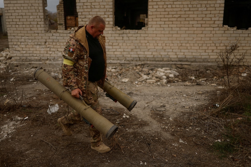 A soldier carries two grenade launchers, one in each hand, past a ruined brick building. 
