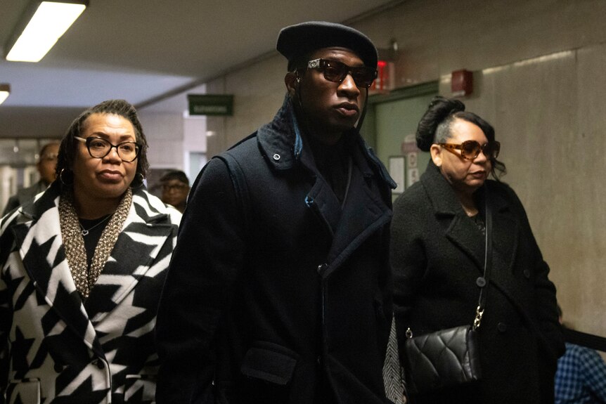 Man in black coat and beret with sunglasses walks in a hallway in front of woman in houndstooth coat. 