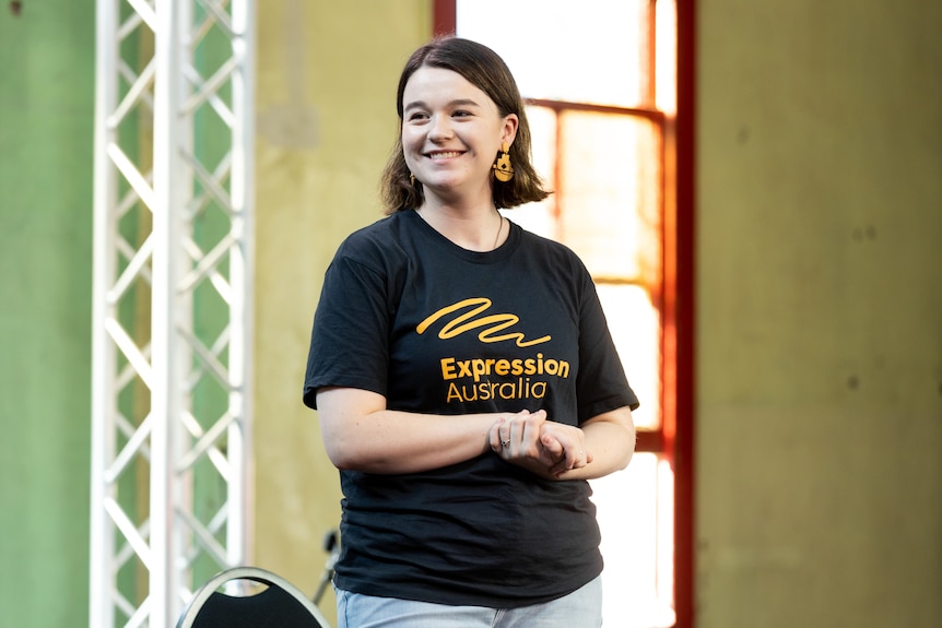 A smiling woman in a black t-shirt with "Expression Australia" on the front, in front of a green background.