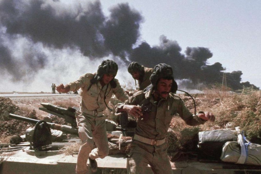 A photograph of men in uniform jumping behind a tank as black smoke billows behind them