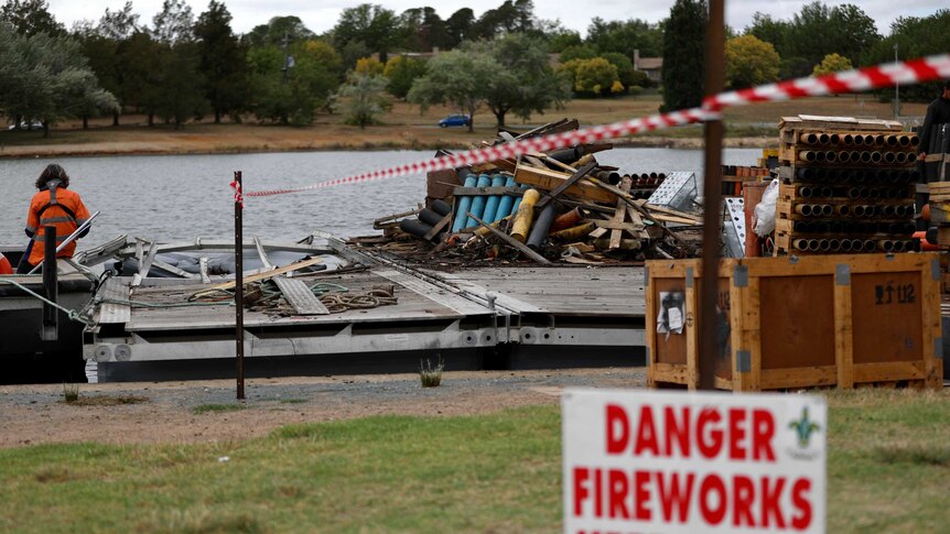 A pile of burnt debris sits on a barge on the water of a lake, next to the shore.