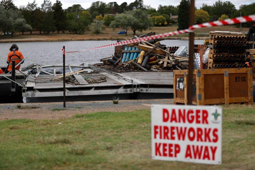 A pile of burnt debris sits on a barge on the water of a lake, next to the shore.