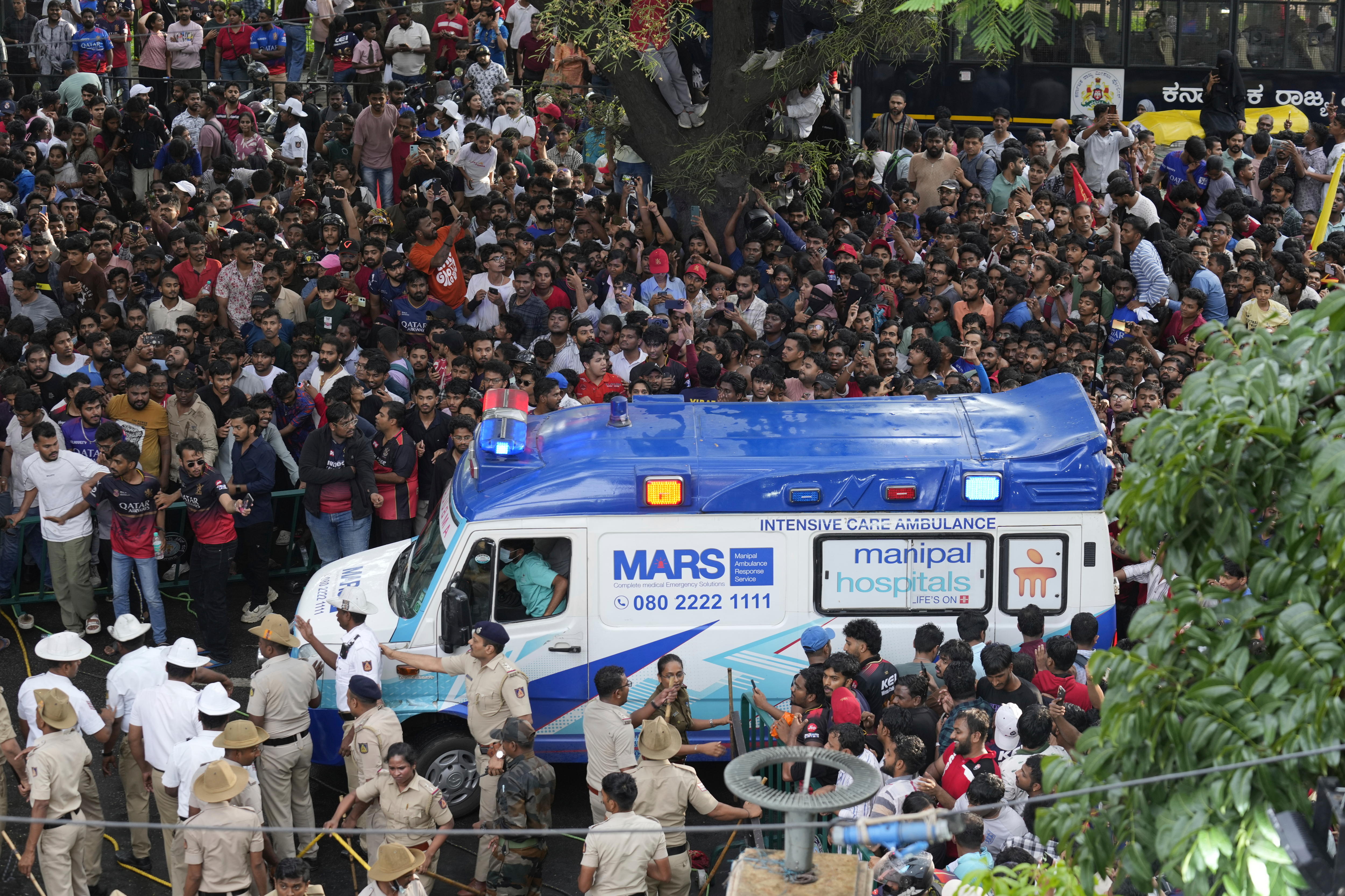 A blue and white ambulance van surrounded by police in tan clothes and a large crowd of people packed around a tree