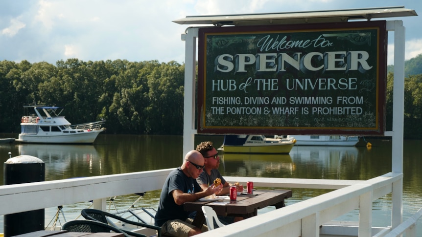 Two men eating lunch by sign that reads Welcome to Spencer Hub of the Universe