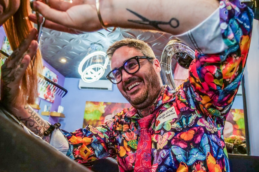 A man wearing a colourful butterfly shirt is cutting a woman's hair in a salon.